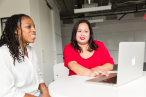 Two women sit in front of a laptop; one uses the mousepad to illustrate something on the screen.